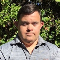 A photograph of Matt Medina. He is wearing a grey monochrome striped shirt with a collar. He has dark, parted hair. He is outside in front of varied green foliage.