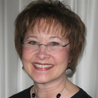 A color portrait of Gail Fanjoy. She is smiling widely with wine red lip color. She has brown hair that is cut into a medium length pixie cut. She is is wearing rimless oval eyeglasses over her blue eyes. She is wearing silver earrings, a black beaded necklace, and a black top.