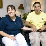 A photograph of a man and a woman, both in wheelchairs, holding hands and smiling in the living room of their home.