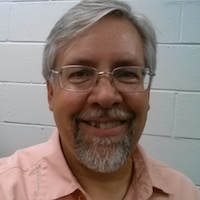 Scott Shepard, a silver-haired man with a similarly silver goatee in a salmon open collared shirt standing against a white cinderblock wall. 