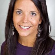 A portrait of Professor Amy Petersen, a broadly smiling woman with dark hair around her shoulders, a purple shirt, against a gray background