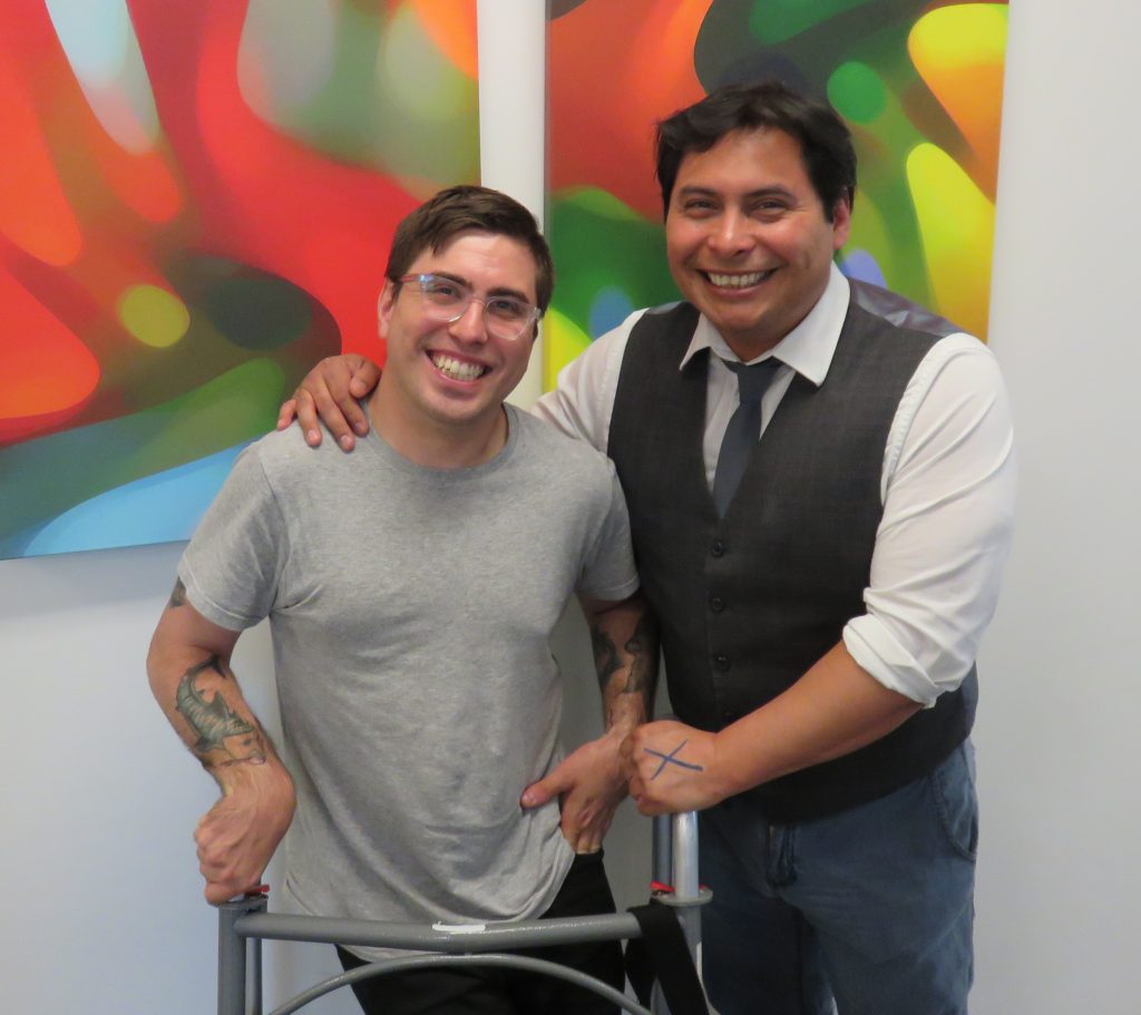 Sean Gray and Edwin Canizalez pose together for a photograph after their Amplified interview. Sean is wearing a gray t-shirt exposing his forearm tattoos. Edwin is wearing a vest and tie and has drawn a straight-edge X on the back of his hand. In the background are two colorful, lumpy-shaped abstract paintings.