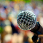 A photograph of a microphone with the metal screen of the microphone head in focus with heavy boke on a very colorful background that hazily looks like a large audience of people.