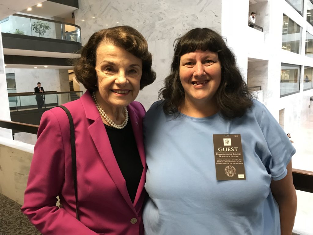 A photograph of Dianne Feinstein and Tia Nelis. THey are in one of the hallways open to the court yard in the white marble Hart Senate Office Building. Tia is wearing her Guest badge.