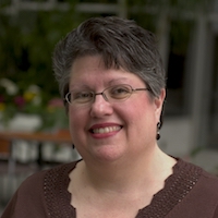 A photograph of Carol Schall, a woman with glasses and short, salt-and-pepper hair. She is wearing a brown shirt with a scalloped collar. The background is a patio with some plants obscured by the depth of field.