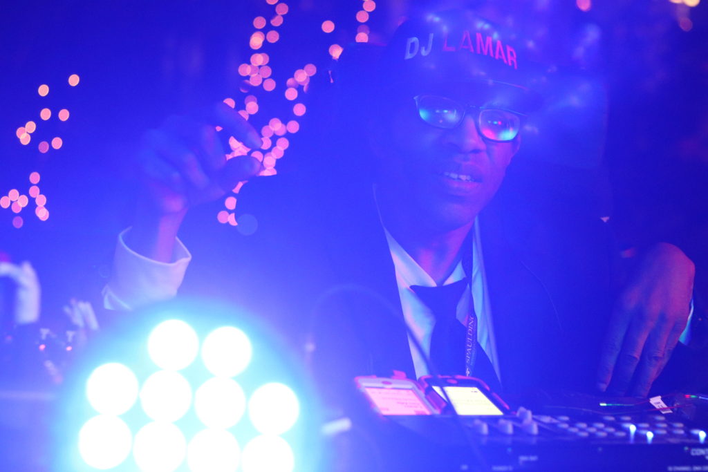 A photograph of DJ Lamar working his lighting and sound mixers during the #TASHbash. One of his lights in the foreground of the image is causing the picture to wash our purple. The blurred Christmas lights behind Lamar look like sparks coming out of his hand.