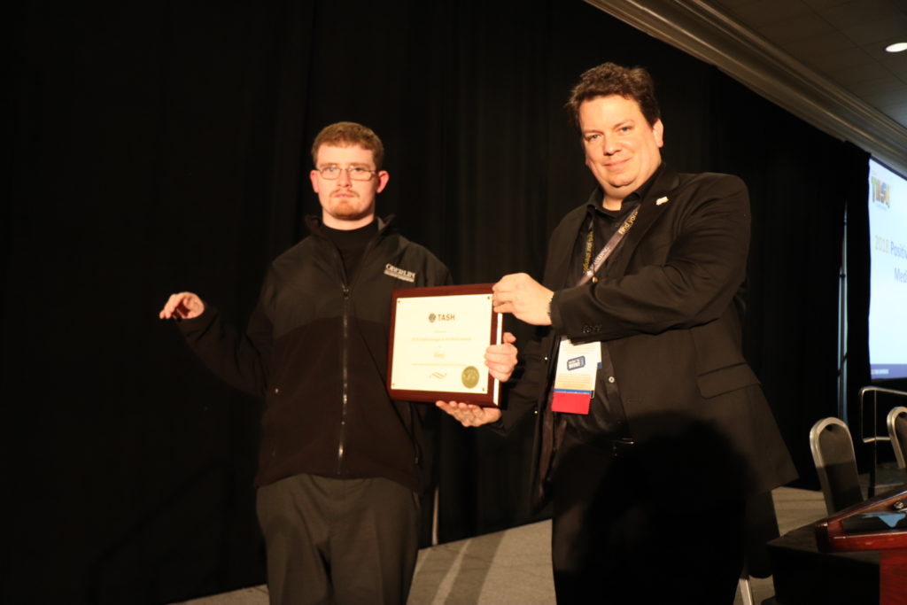 A photograph of David Savarese accepting the Positive Images in the Media Award. TASH staff member Donald Taylor hands him a plaque on a black curtained stage.