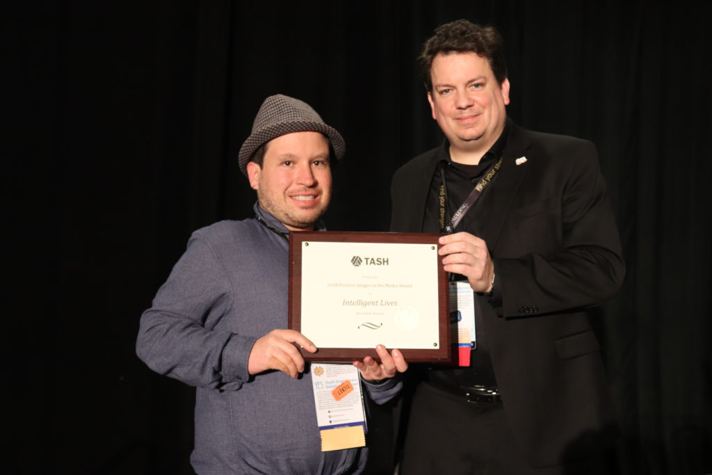 A photograph of Micah Fialka-Feldman accepting the Positive Images in the Media Award. TASH staff member Donald Taylor hands him a plaque on a black curtained stage.