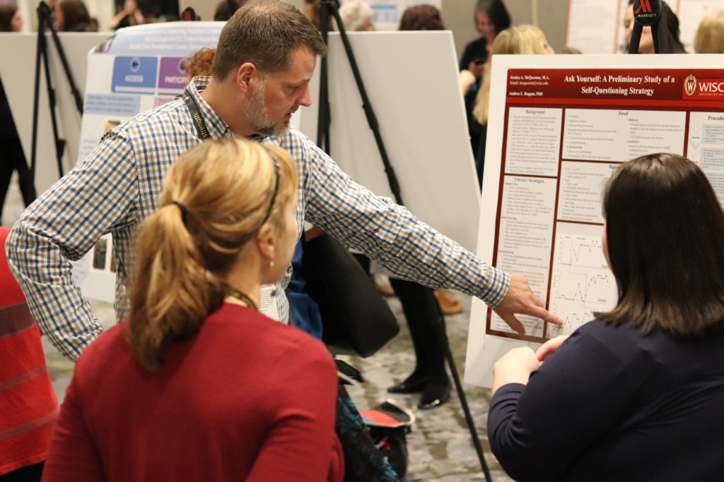 A photograph of Erik Carter pointing to a set of graphs on a poster while one of the poster authors and another discuss the study.