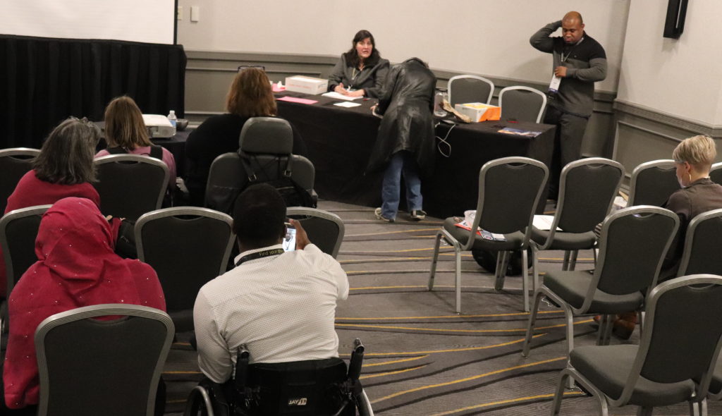 A photograph of the Self-Advocacy Forum: a group of people in audience seating with some of them at the front of the room working on a variety of papers.
