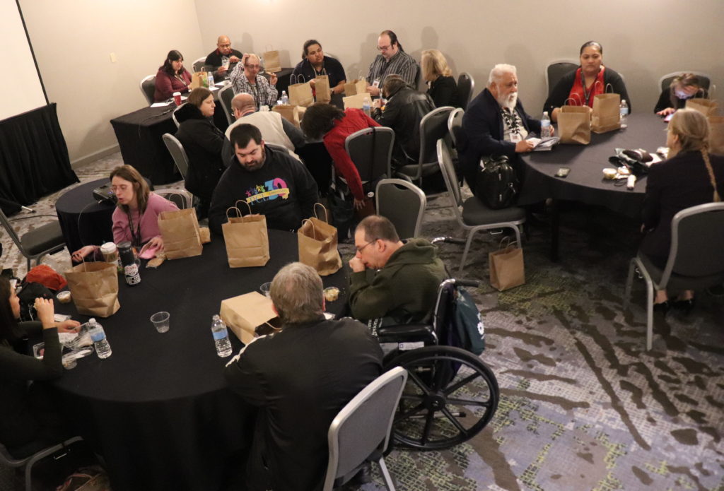 A photograph of the Self-Advocacy Let's Connect Luncheon. A medium-sized group of people sitting at round tables in a small conference room.