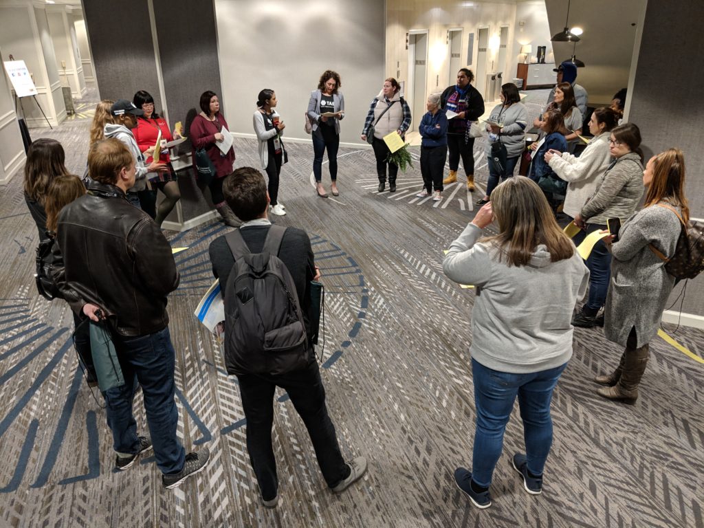 A photograph of the Conference Volunteers orientation: a large group of people standing in a large circle, all referring to a packet of handouts.
