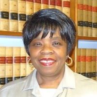 A photograph of Barbara Ransom, an African-American woman smiling with narrow eyes, short black hair with silver streaks parted with bangs. She is wearing a white shirt with a collar and a beige jacket. In the background is a large shelf of pale yellow law books with red and black stripes on the spines.