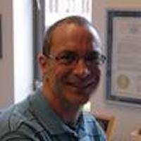 A low-resolution photograph of Keith Hyatt. It is quarter profile and he is turning his head towards the viewer. He has tightly cropped hair, a significant widow's peak hairline and glasses. He is smiling broadly. Some diplomas are visible on the wall behind him.