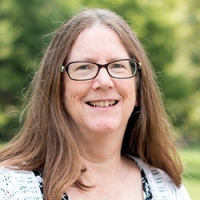 A portrait of Lou-Ann Land. She has glasses and shoulder-length brown hair parted in the middle. Se is outside, standing against a leafy tree that is obscured by the depth of field.