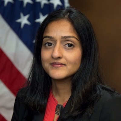 A portrait of Vanita Gupta, a South Asian woman with shoulder-length dark hair parted slightly to her left. It is an official DOJ portrait, so there is a brown wood-panel wall and a U.S. flag in the background.