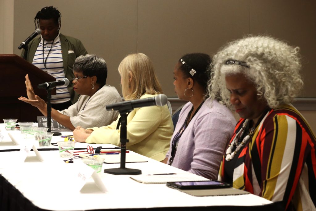 A photograph looking down the table of panelists with Barbara Ransom at the end gesturing as she speaks and moderator Tanesha Williams looking on, standing at the podium.
