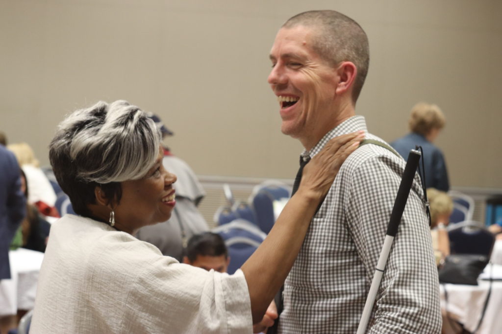A photograph of Barbara Ransom and a man with a white stick. Barbara reaches out to put her hand on the man's shoulder as they both smile broadly.