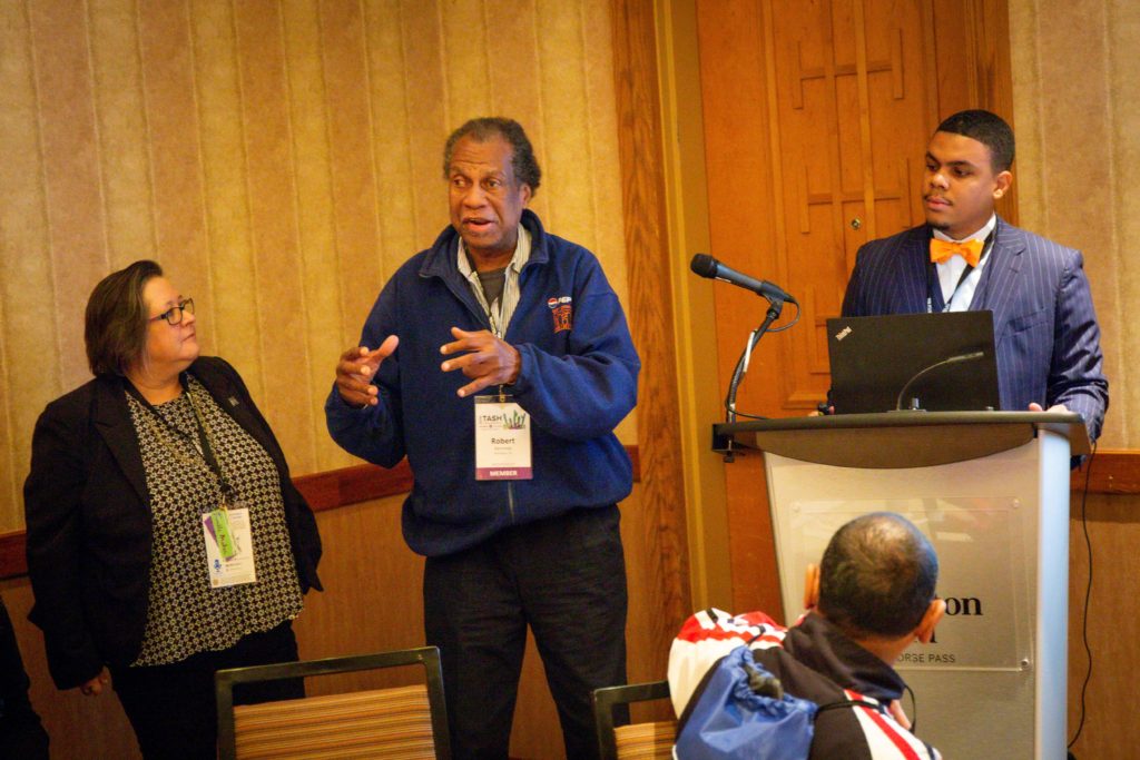 A photograph of a group presentation. One person in a bowtie stands behind a podium watching as a man in a windbreaker gestures while talking.