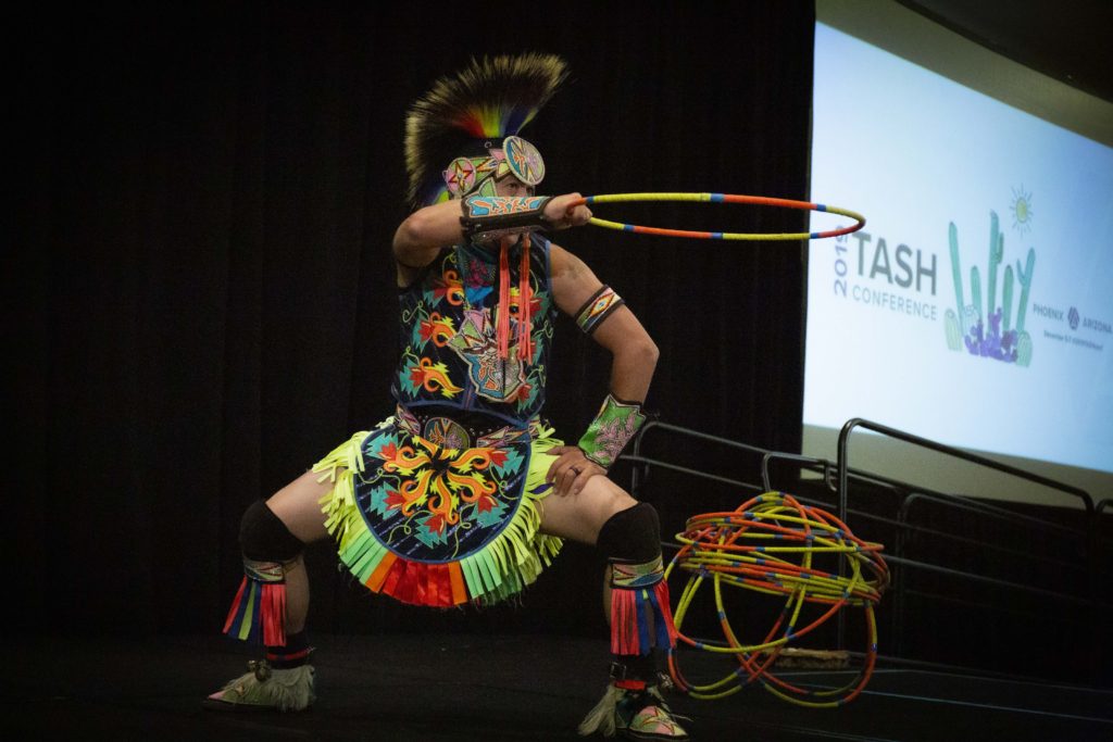 A photograph of Brian Hammill in a colorful tribal outfit holding the climactic pose of his dance: in a squat with one hand on one knee and holding one of his hoops straight out at eye level.