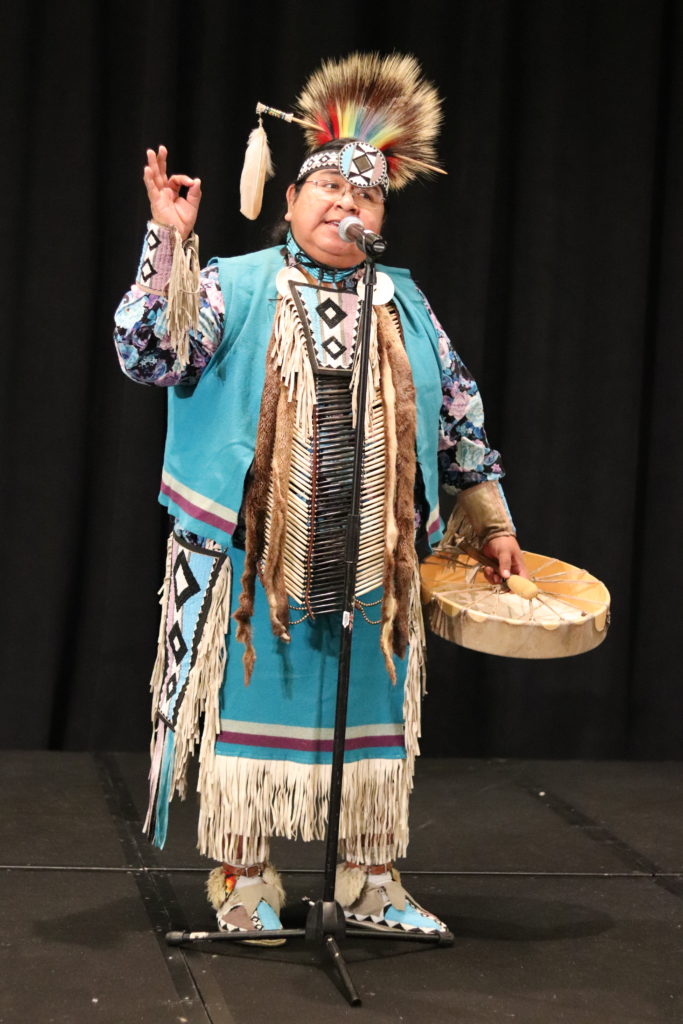 A photograph of the drummer for Native Spirit Productions. He is gesturing with his hand up to emphasize a point as he speaks into a microphone. He is wearing a turquoise native american outfit and holding a hand drum at his side.