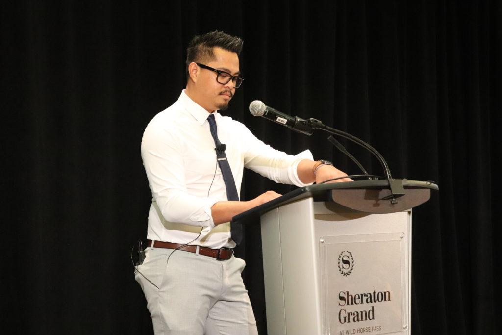 Photograph of Raymond Guron. He has a black, spiky pompadour and glasses. He is wearing a white shirt, tie and khaki pants. He is standing at a podium with a microphone.