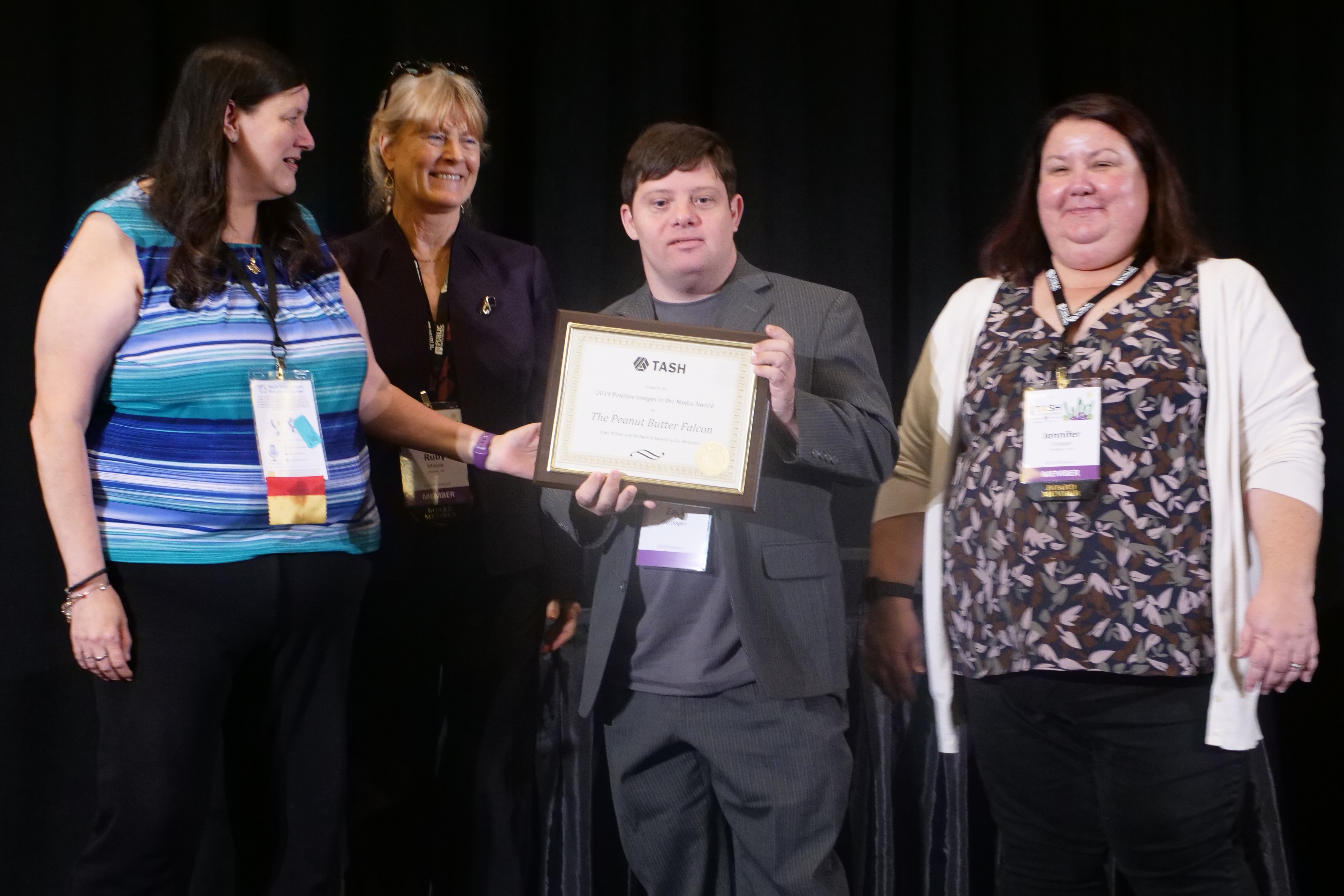 A photograph of Zack Gottsagen accepting the Positive Images in the Media Award from Tia Nelis. Ruby Moore and Jenny Lengyel are also in the group.