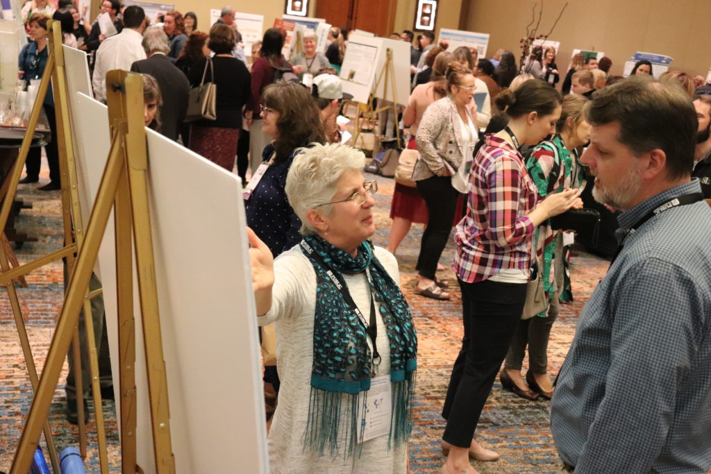 A photograph of the Poster Presentation hall. Two people discuss a poster in the foreground; in the background is a full, busy room of posters.