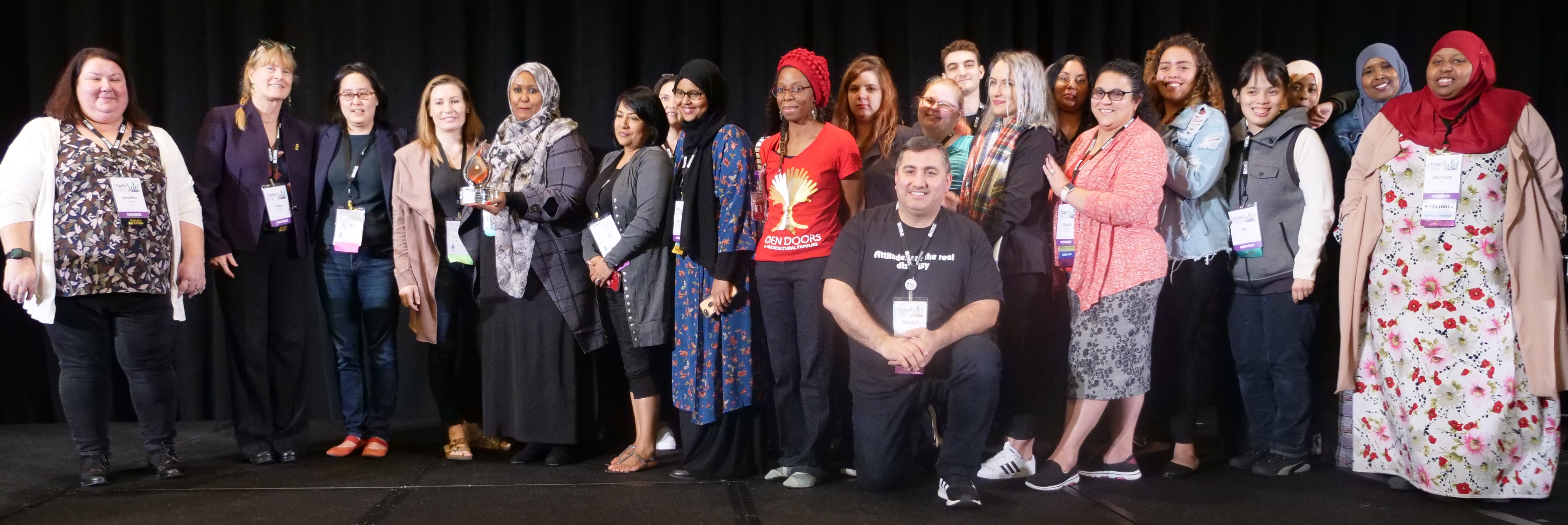 A photograph of the Open Doors for Multicultural Families delegation accepting the Ralph Edwards Diversity and Social Impact Award for Ginger Kwan. A large, diverse group of people wearing casual and professional outfits and head-coverings from multiple cultures.