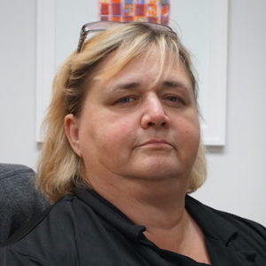 A portrait of Cheri Mitchell. Her shoulders are turned 3/4 and she is looking slightly left of straight-on. She is wearing a black collared shirt and her glasses up on her head. You can see the back of her power chair and a paining in the background.