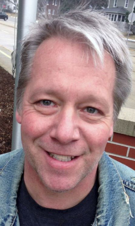 A photograph of Christopher Kliewer. He has a shock of silver hair leaning to one side and is wearing a denim jacket and black t-shirt. He is out on the street in front of a brick half-wall.