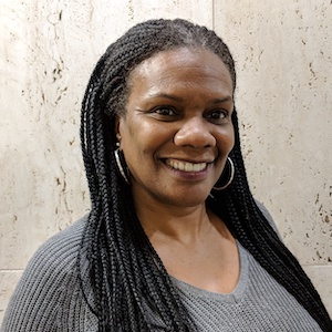 A portrait of Sheila Harrison. She is a black woman turned a quarter to her left. She has long small braids over each shoulder and is wearing a v-neck grey ribbed shirt. She is standing against a pale marble panel wall.