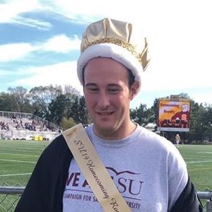 A photograph of Will Fried at a school event. He is wearing a large gold cloth crown and a sash. In the background is a school sports field and bleachers.