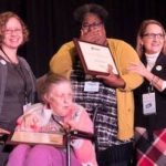 A photograph of an African-American woman holding a plaque with her hand over her mouth in surprise and tearful joy, standing behind a white woman in a wheelchair. The TASH presenters stand to their left and right.