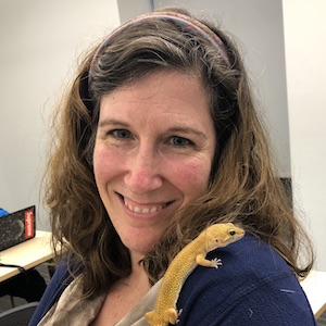 A photograph of Deborah Taub. She has shoulder-length brown hair and a headband. She is smiling. There is a yellow lizard on her shoulder.
