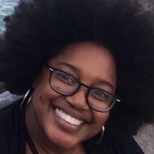 A portrait of LaQuita Montgomery. She is an African American woman. The photograph is close-in and she is tilting her head to one side. Her hair fills most of the frame. She has glasses and is smiling broadly.