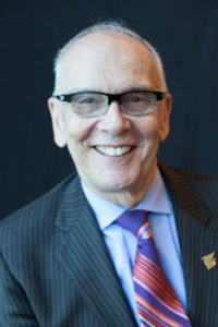 A portrait of Curt Decker. He has a thin high hairline and horn-rimmed glasses. He is wearing a dark striped suite and a colorful tie in pink and purple stripes.