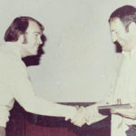 An old, slightly damaged photograph of Wayne Sailor and Lou Brown smiling at one another and shaking hands in front of a projector screen. Lou has a plaque and gavel that he is presenting to Wayne.