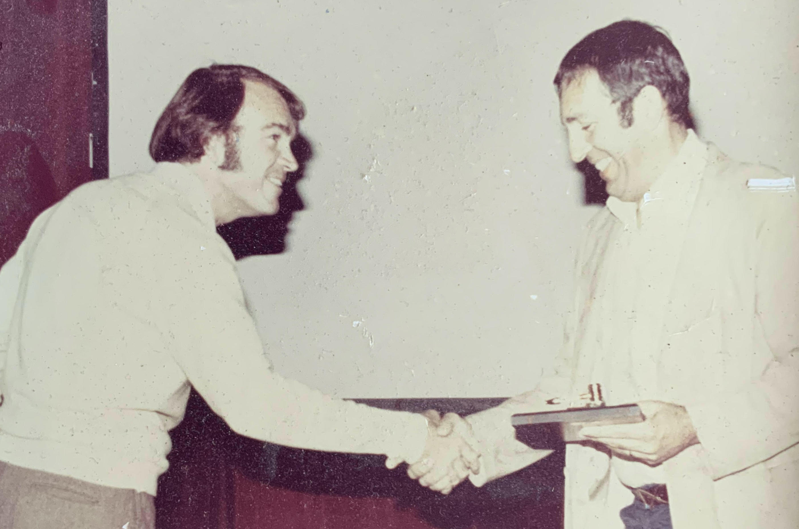 An old, slightly damaged photograph of Wayne Sailor and Lou Brown smiling at one another and shaking hands in front of a projector screen. Lou has a plaque and gavel that he is presenting to Wayne.