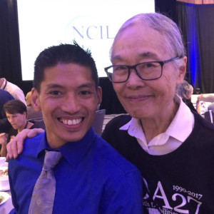 A photograph of Mathew McCollough with Yoshiko Dart. They are standing quarter profile with Yoshiko slightly behind Mathew. Mathew has close cropped black hair a large grin and is wearing a royal blue shirt with a tie.