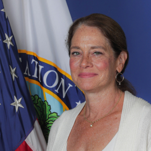 Official government portrait of Katherine Neas. She has dark hair pulled back into a ponytail. She is wearing a ribbed, cream-colored shirt. Behind her are the United States flag and a flag with the seal of the Department of Education