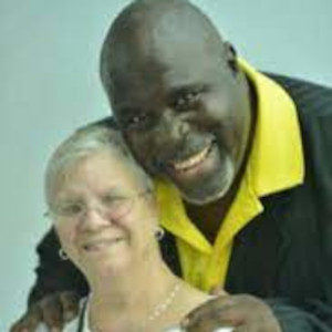 A photograph of Ricardo and Donna Thornton. Ricardo is a black man with a shaved head and a large smile. Donna is a white woman with grey hair and glasses. Ricardo leans into the frame and rests his hands on Donna's shoulders.