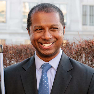 A portrait of Sachin Dev Pavithran. He has a crooked smile and is wearing a dark suite. The hedgerow and marble of a government building are behind him. The handle end of his white cane is slightly in frame.