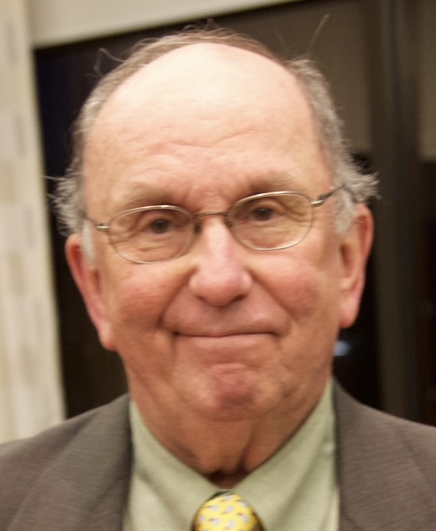A portrait of Michael Lottman. He has silver-rimmed glasses and a lopsided smile. He is wearing a brown suite, pale green shirt and a yellow tie.