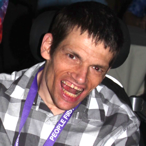 A color photograph of Ryan Duncanwood. He looks extremely excited and is smiling. Some of the supports of his power chair are visible behind him and he is wearing a purple lanyard that reads People First.