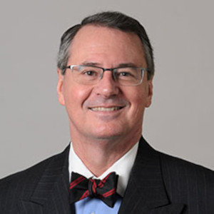 A portrait of Craig Kennedy. He has parted silver and black hair, glasses, a two-toned collared shirt under a navy blazer with a dark striped bow tie.