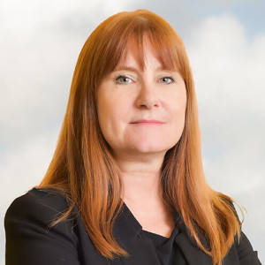 A portrait of Eve Hill, JD. She has long orange hair and bangs. She is wearing a black suite and is against a cloudy sky.