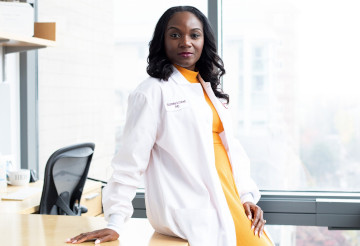 A photograph of Kizzmekia Corbett. She is a black woman with high, parted hair, wearing a bright yellow dress and a white lab coat. She is leaning against a desk in an office.