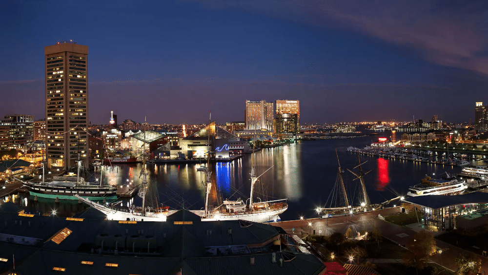 A photograph of a nighttime view of the Inner Harbor from the Hyatt Regency. Numerous boats are illuminated by dock lights and the lights of the city spread into the distance.