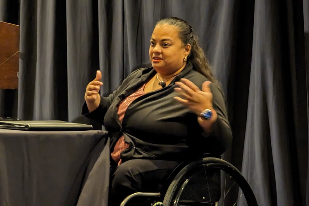 A photograph of Anjali-Forber-Pratt on stage with a black backdrop, gesturing with both hands as she addresses the Conference general session.
